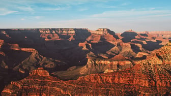 Sunrise light bathes the layered red-rock buttes and ridgelines of the Grand Canyon under a clear blue sky, with deep shadows filling the canyon's side gorges, Arizona.